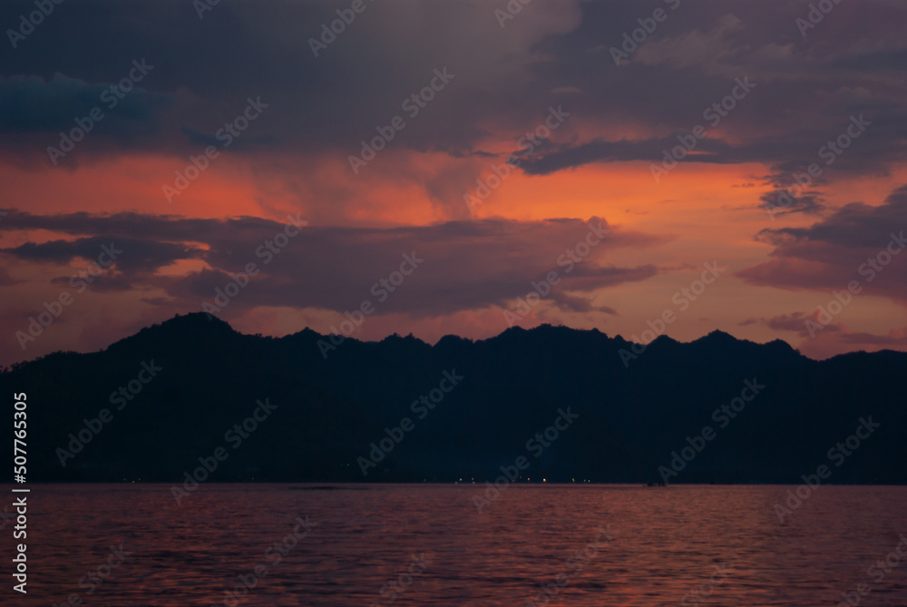 Dark majestic purple sunset on ocean with dark silhouette mountains as line, orange, pink saturated reflection sunbeams in calm water. Dramatis indonesian landscape in evening, beautiful trip in asia.