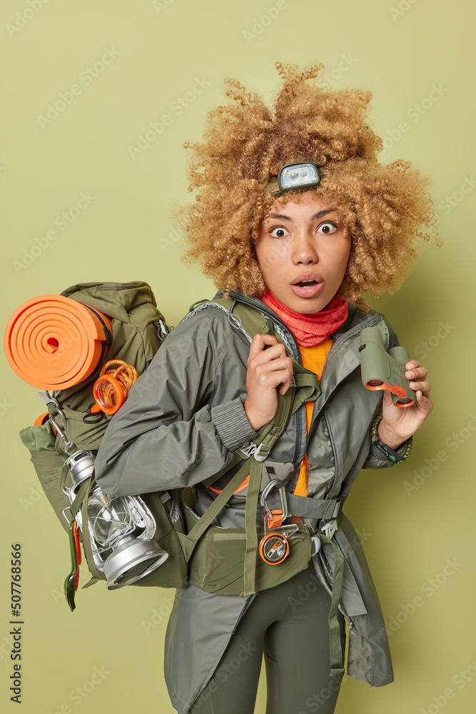 Vertical shot of impressed curly haired woman dressed in activewear holds binoculars stares shocked at camera dressed in comfortale clothes for hiking carries rucksack isolated over green background
