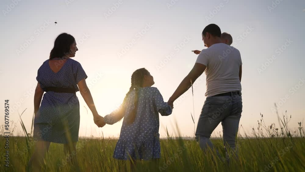 Happy family. Family walk in the meadow. Park with green grass. Parents hold their children by the hands in nature. Natural park at sunset. Active lifestyle. Cheerful happy family in the park outdoors Happy family. Family walk in the meadow. Park with green grass. Parents hold their children by the hands in nature. Natural park at sunset. Active lifestyle. Cheerful happy family in the park outdoors