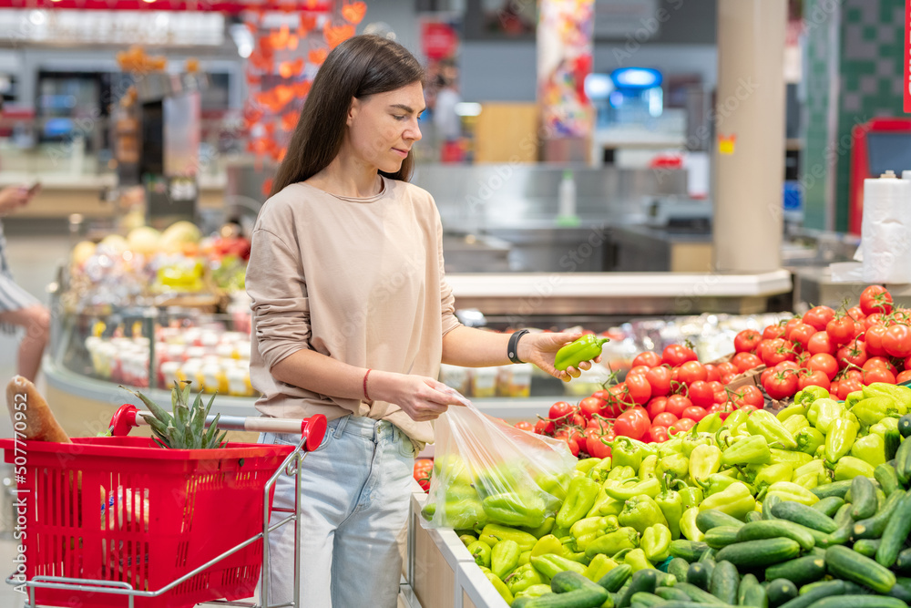 Portrait of young adult woman standing in supermarket with shopping cart choosing sweet peppers Portrait of young adult woman standing in supermarket with shopping cart choosing sweet peppers