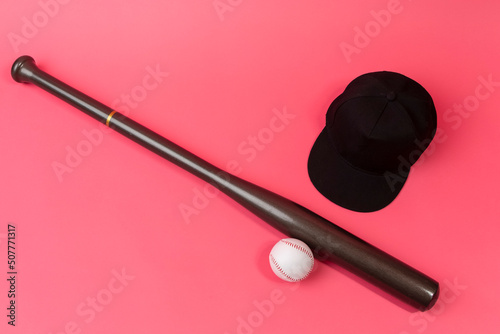 Image of Stylish Laquered Wooden Brown American Baseball Bat Placed With Clean Leather White Baseball Ball and Black Cap Over Trendy Coral Pink Background.