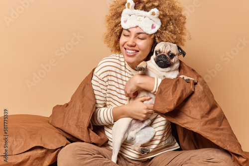 Positive young woman pet owner embraces pug dog with love stays in bed after awakening dressed in comfortable pajama covered with blanket isolated over beige background. Domestic animals concept