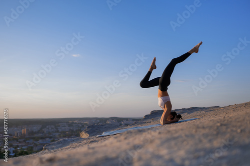Beautiful young caucasian girl practices yoga and performs a headstand with her legs crossed on a rock on a background of sunset and cityscape © Myron Muza