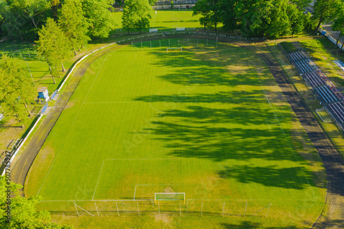 Prowincjonalny stadion. W centrum porośnięte murawą boisko piłkarskie, wokół bieżnia i niewielka trybuna dla kibiców. Widok z drona.