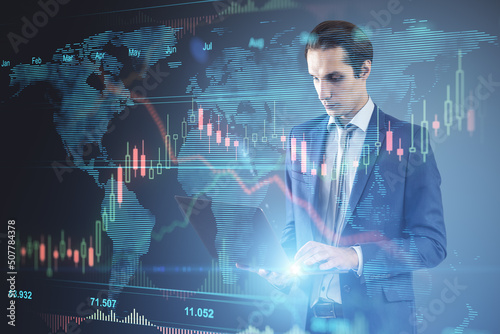 Attractive young european businessman using laptop with glowing forex chart hologram on blurry background. Finance  trade and fintech concept. Double exposure.