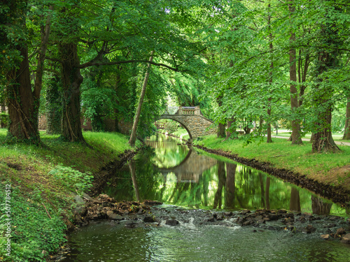 Park dworski w mieście Iłowa w Polsce. Płynąca leniwie przez park rzeka. Nad rzeką zabytkowy, kamienny mostek. Na brzegu zielona trawa, wokół drzewa.