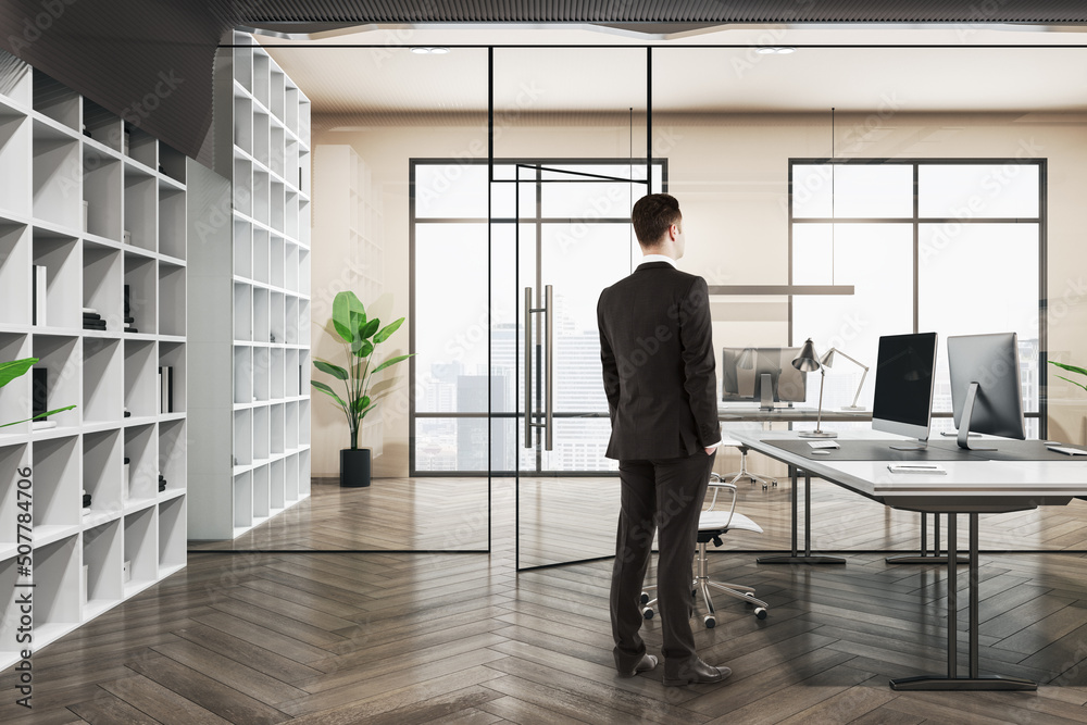 Fototapeta premium Attractive thoughtful european businessman standing in wooden and concrete office interior with furnituire and window with city view. CEO, executive and consulting concept.