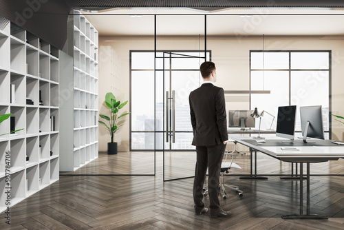 Attractive thoughtful european businessman standing in wooden and concrete office interior with furnituire and window with city view. CEO, executive and consulting concept.