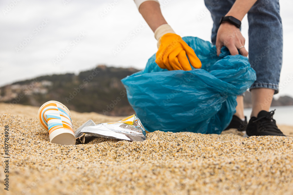 Obraz premium A woman in gloves with a special blue bag picks up garbage among the sand along the coast. The problem of environmental pollution. Cleaning up trash on the beach