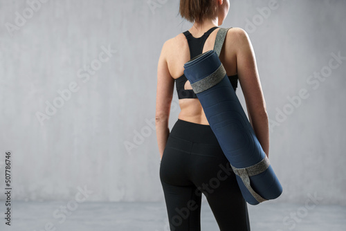 Rear view of sports woman in fashion black sport clothes carrying fitness mat in gym, over gray background