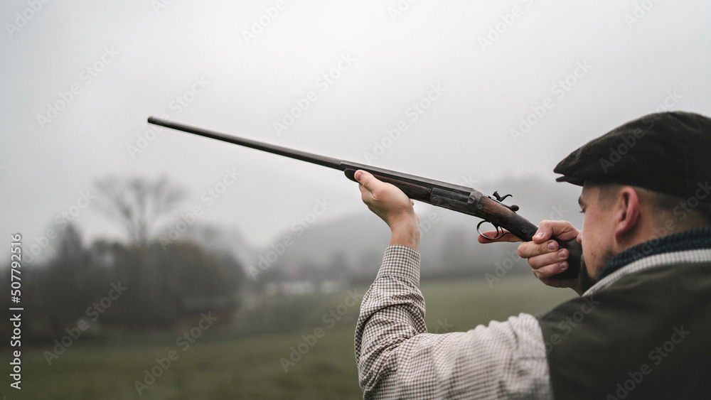 Hunter man in traditional shooting clothes on field aiming with shotgun