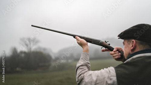 Fotografie, Obraz Hunter man in traditional shooting clothes on field aiming with shotgun