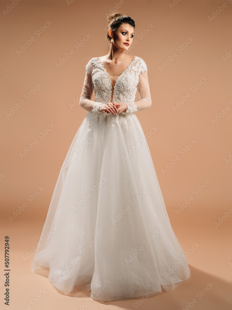 Brunette bride in white wedding dress with deep decollete smiling and looking in camera. Lace bridal gown with transparent long sleeves, front slit, tulle skirt and floral embroidery. Happy marriage.