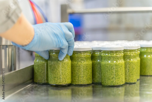Pesto sauce with basil Industrial process. Many jars of sauce prepared for labeling. Worker at a food factory selects jars of pesto sauce by quality, sorts, separates closed lids
