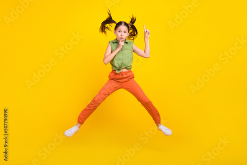 Photo of shiny serious student girl wear green shirt smiling jumping high finger lips asking keeping silence isolated yellow color background