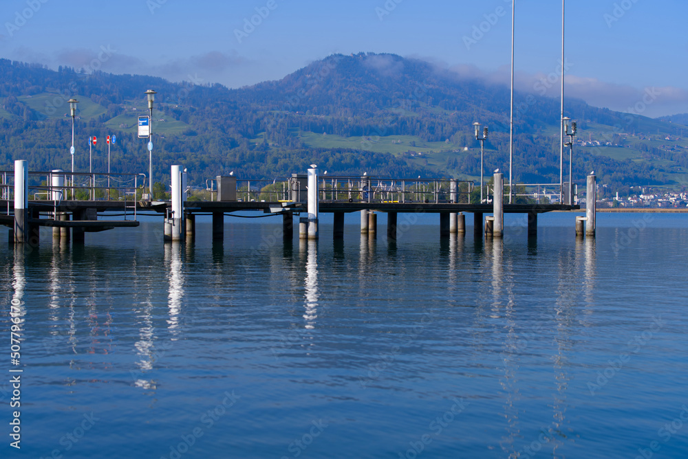 Fototapeta premium Scenic landscape with Lake Zürich and pier with flags seen from City of Rapperswil-Jona on a sunny spring day. Photo taken April 28th, 2022, Rapperswil-Jona, Switzerland.