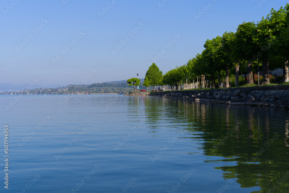 Waterfront with boardwalk and tree alley at border of Lake Zürich at City of Rapperswil-Jona on a sunny spring day. Photo taken April 28th, 2022, Rapperswil-Jona, Switzerland.