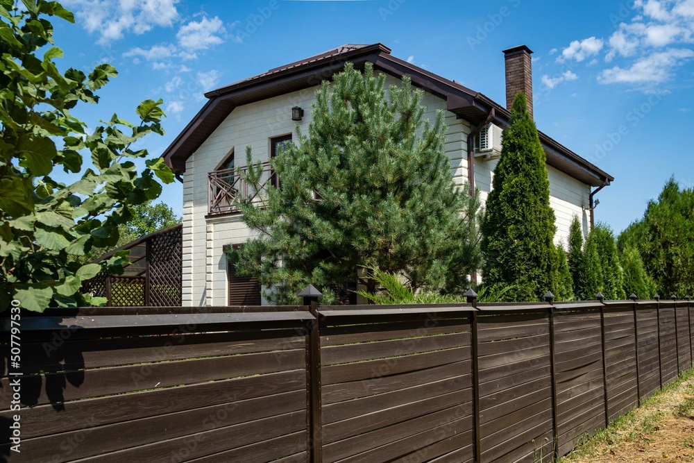 Krasnodar, Russia - Aug 10, 2020: Hedge of evergreen trees behind brown wooden fence with two-story country house. Natural defense against harmful odors and disease-causing bacteria and viruses.