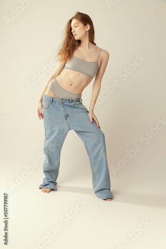 Full-length portrait of slim beautiful young girl posing in grey underwear and oversized jeans isolated over grey studio background. Weight-loss