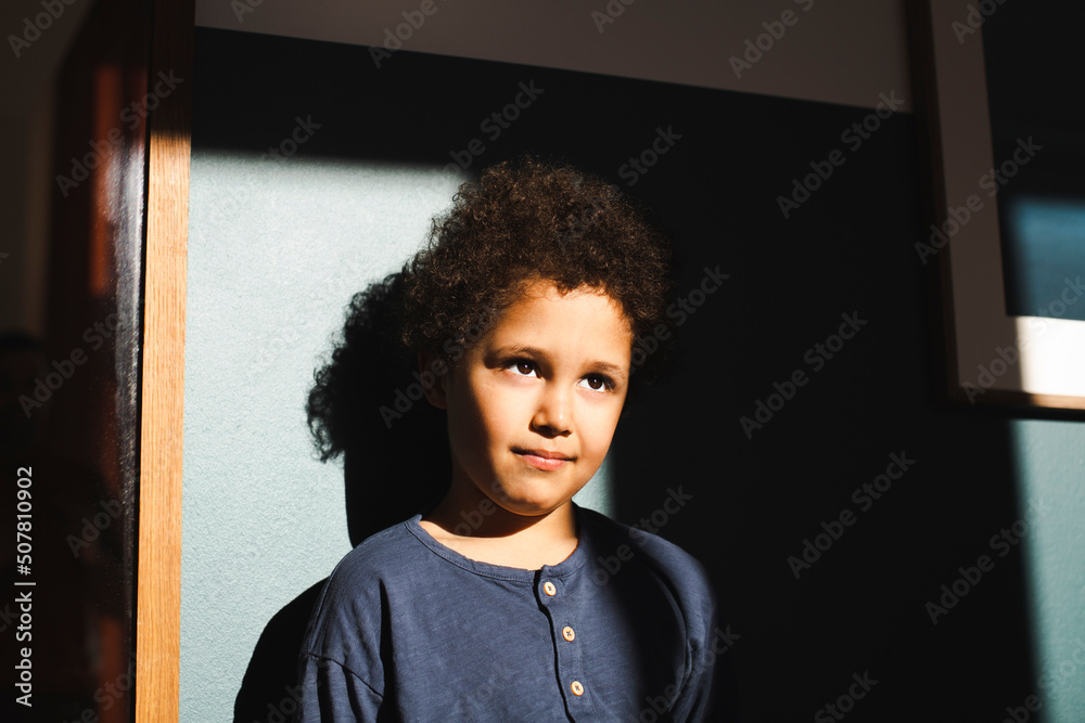 Boy looking away standing in sunlight against wall at home Boy looking away standing in sunlight against wall at home