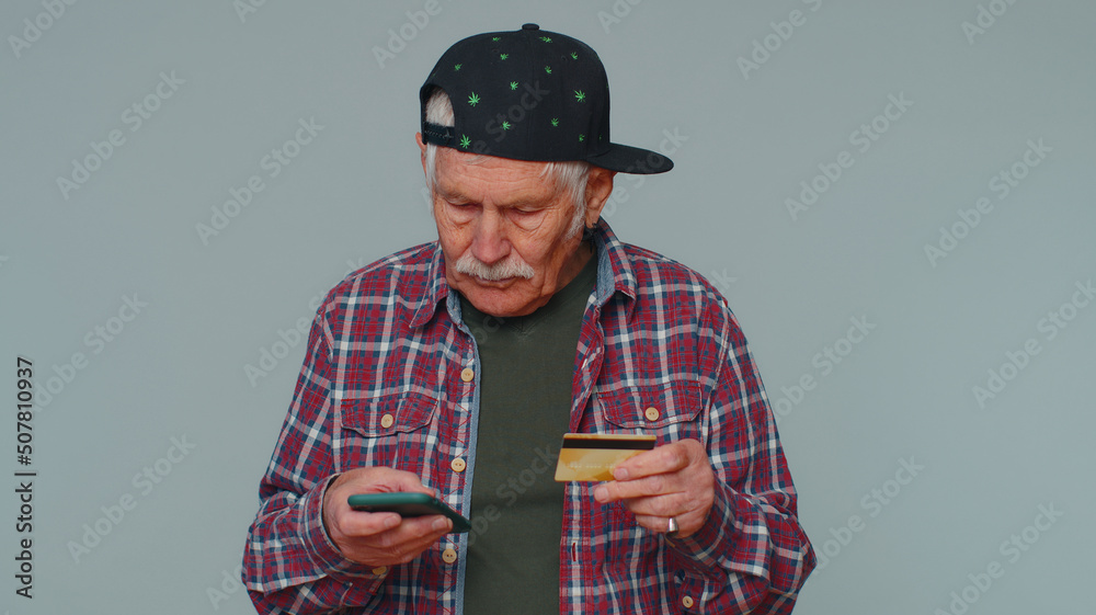 Sincere mature man customer using credit bank card and smartphone while transferring money, purchases online shopping, payment. Finance and internet. Senior grandfather alone on gray wall background Sincere mature man customer using credit bank card and smartphone while transferring money, purchases online shopping, payment. Finance and internet. Senior grandfather alone on gray wall background