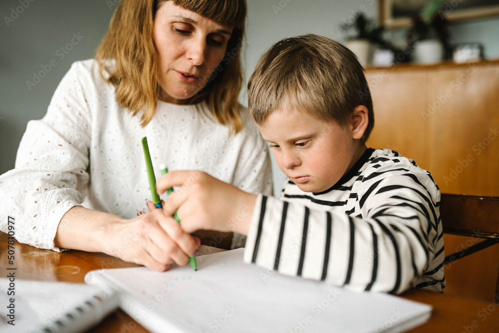Boy with disability taking support of mother while drawing in book at home Boy with disability taking support of mother while drawing in book at home