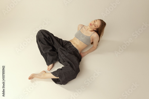 Portrait of young slim girl in top and giant black jeans leying on floor, posing isolated over grey studio background. Weight-loss concept photo