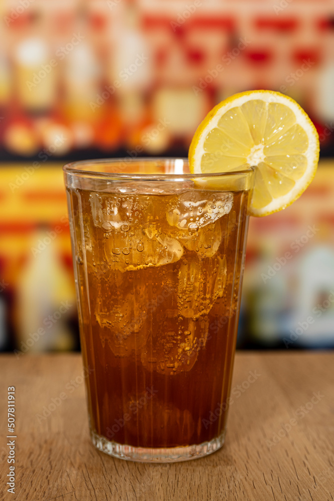 Glass with sparkling cola, ice and lemon on a wooden counter in front of a bar. Glass with sparkling cola, ice and lemon on a wooden counter in front of a bar.