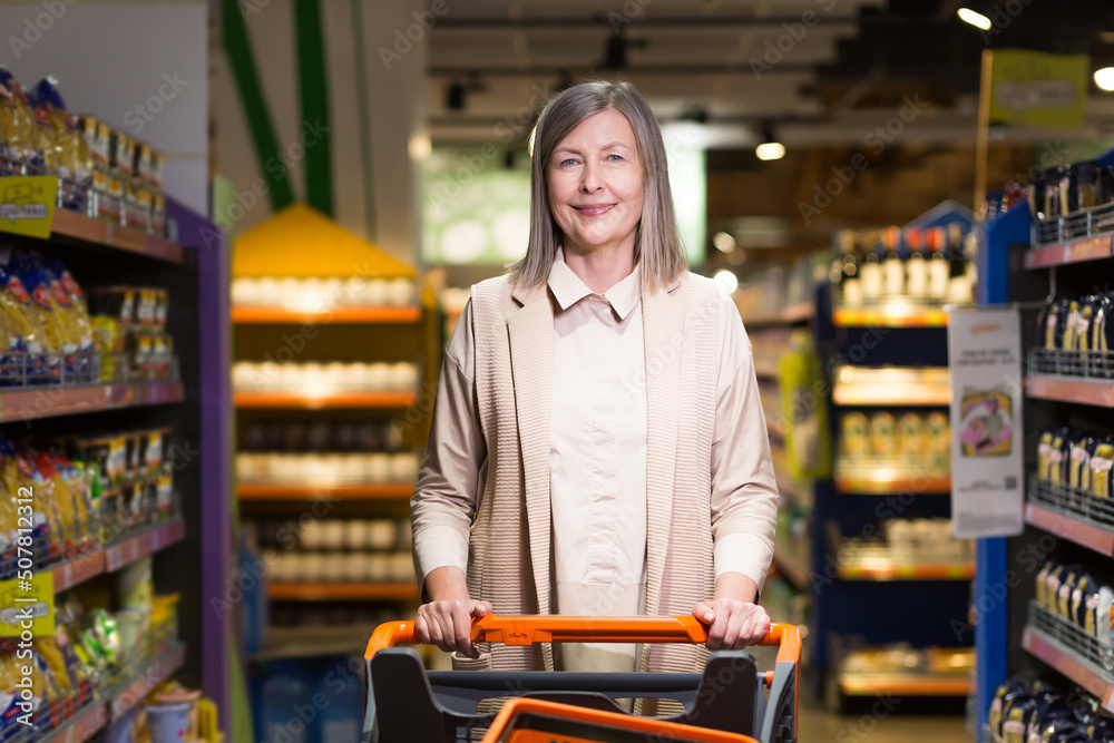 Portrait of a beautiful senior woman in a store, supermarket. He stands, holds a food cart in his hands, looks at the camera, smiles Portrait of a beautiful senior woman in a store, supermarket. He stands, holds a food cart in his hands, looks at the camera, smiles