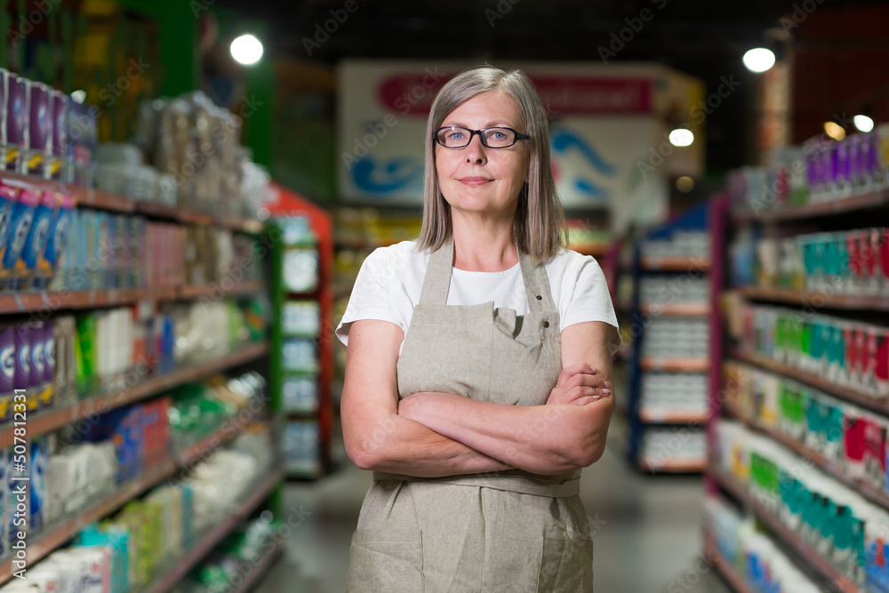 Portrait of senior woman in glasses manager of grocery store, supermarket. Standing in work clothes, arms crossed, looking at the camera, smiling