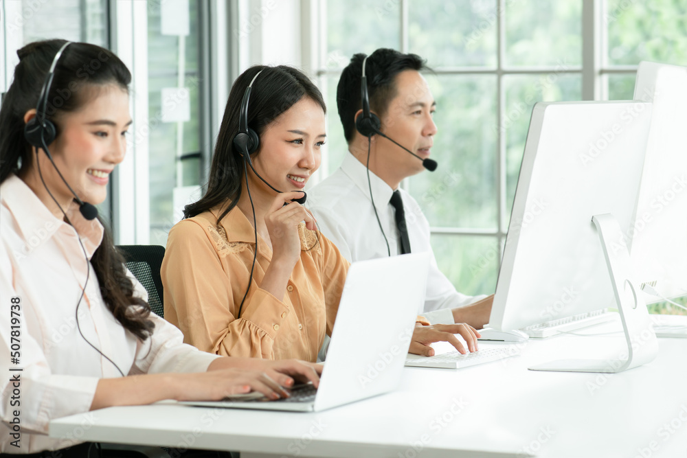 Selective focus on young operator woman touching microphone on headset and working with smiling and doing customer support at work. Group of Asian employee work in telemarketing customer service teams