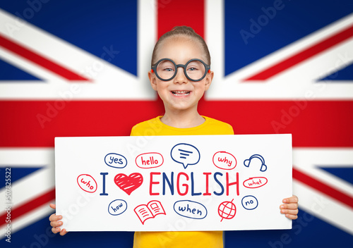 Beautiful cute little girl holding a white board with i love English text and illustrations in front of blurry flag of United Kingdom. photo