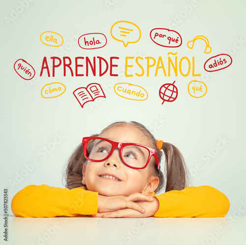 Beautiful cute little girl with eyeglasses looking at Learn Spanish text in Spanish and illustrations. English: Learn Spanish. Foreign language learning concept.
