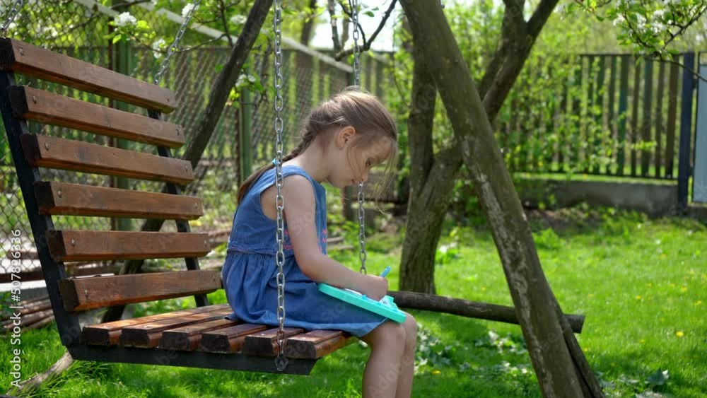 Beautiful little girl is learning to draw while sitting on a swing on a sunny day. Side view Beautiful little girl is learning to draw while sitting on a swing on a sunny day. Side view