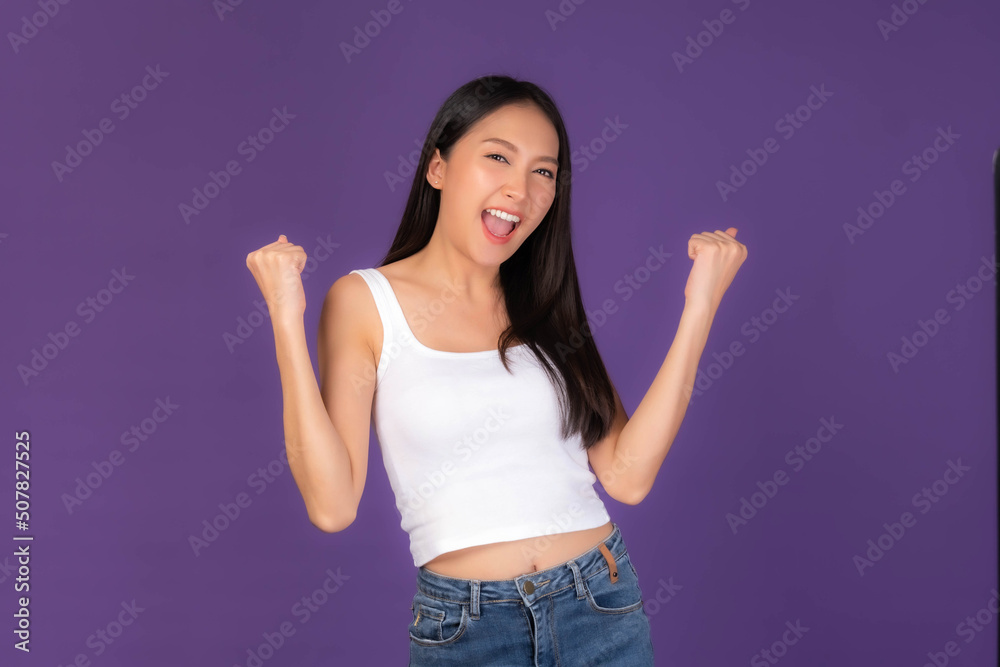Portrait of excited Beautiful Asian brunette woman cute girl in white tank top raising hand in the air laughing cheerfully gesturing for winner , smiling , look camera isolated purple background Portrait of excited Beautiful Asian brunette woman cute girl in white tank top raising hand in the air laughing cheerfully gesturing for winner , smiling , look camera isolated purple background