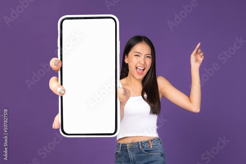 Full body length photo portrait of beautiful Asian young woman ,Excited surprised girl showing big smart phone with blank screen , white screen isolated on purple background , Mock Up Image