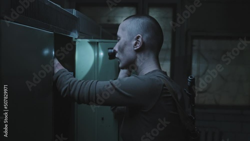 Medium tracking of bruised female soldier with shaved head holding flashlight, looting in abandoned dark locker room photo