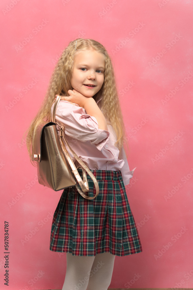 portrait of a little girl with a backpack. schoolgirl in uniform. pink background.