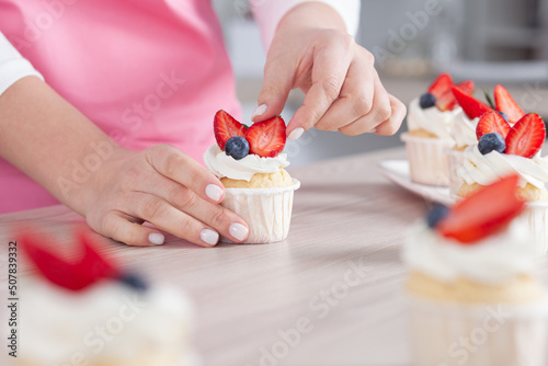 Pastry chef confectioner young caucasian woman in the kitchen. She is decorating a cupcake with with strawberries and blueberries