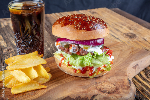 Juicy and appetizing burger with tomatoes, lettuce, cucumber, melted cheese and onions with brioche bun served on a wooden board with fries