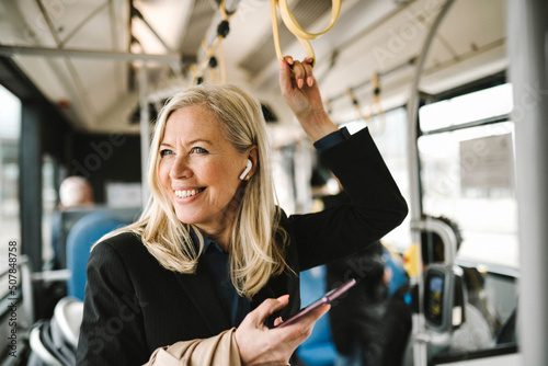Happy businesswoman talking on smart phone holding grab handle while commuting through bus