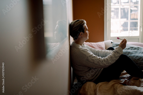 Side view of upset woman holding pregnancy test result sitting on bed at home photo