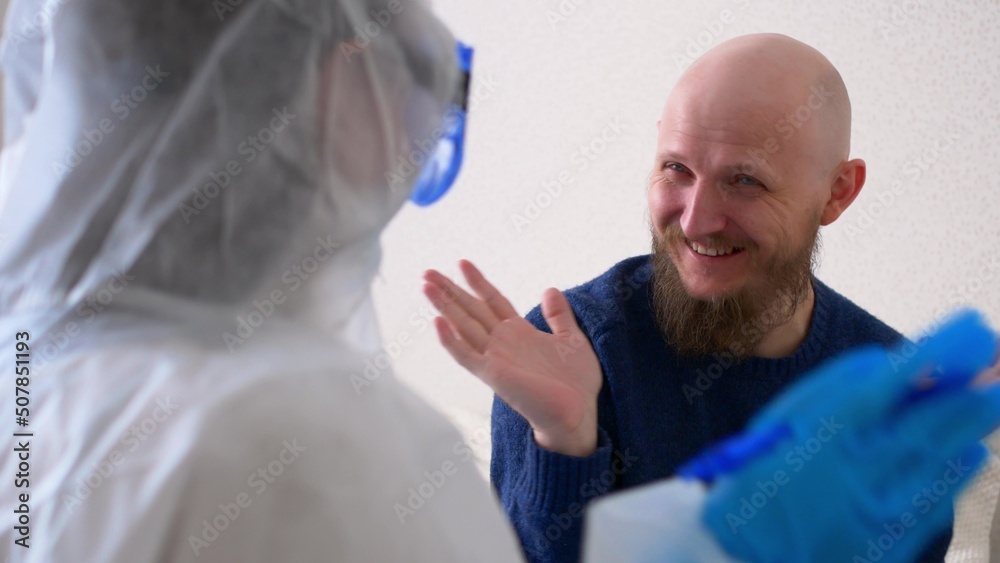 The doctor came to the patient and amuses him in order to cheer up the patient. A doctor in a mask and protective suit is fooling around with a patient, a middle-aged man with a beard. The doctor came to the patient and amuses him in order to cheer up the patient. A doctor in a mask and protective suit is fooling around with a patient, a middle-aged man with a beard.