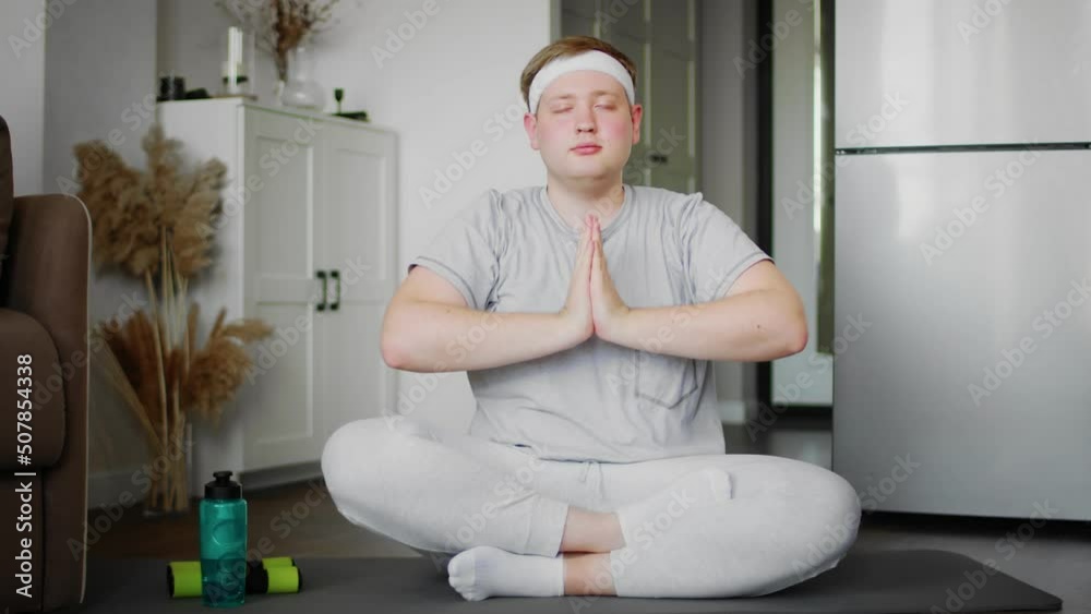 The guy practices yoga sitting on the floor in the lotus position at home in the living room. Relaxation and meditation alone. The guy practices yoga sitting on the floor in the lotus position at home in the living room. Relaxation and meditation alone.