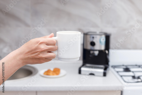 cup of coffee in hand background of a coffee machine and a croissant in the kitchen. coffee break