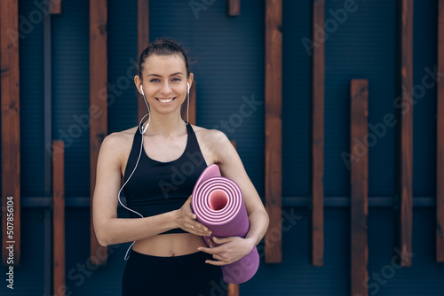 Girl standing, smiling, holding yoga mat.