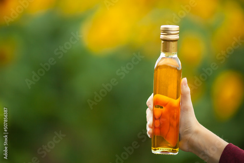 Female hand holding a glass bottle of vegetable oil on a blurred green and yellow background. Sunflower, sesame, linseed or corn oil. Concept of natural healthy product. Copy space. Selective focus.