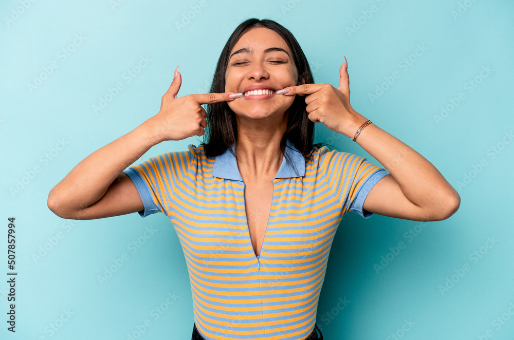 Fototapeta premium Young hispanic woman isolated on blue background smiles, pointing fingers at mouth.