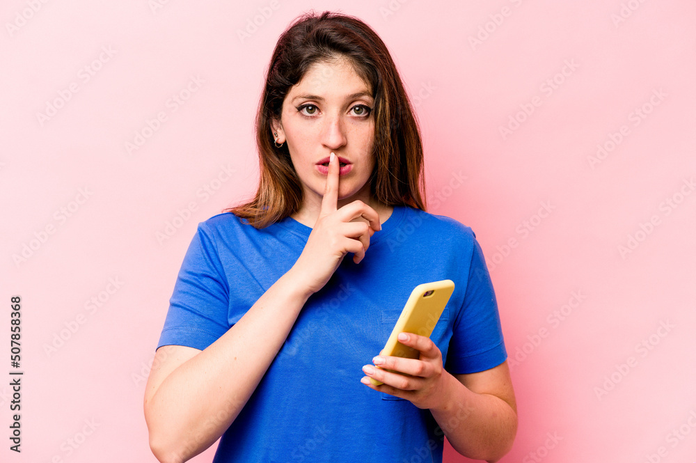 Young caucasian woman holding mobile phone isolated on pink background keeping a secret or asking for silence. Young caucasian woman holding mobile phone isolated on pink background keeping a secret or asking for silence.