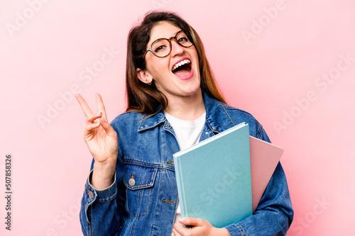 Young student caucasian woman isolated on pink background joyful and carefree showing a peace symbol with fingers.